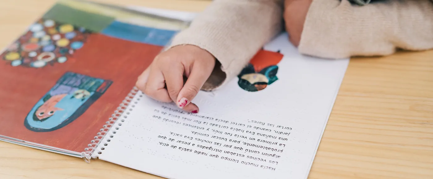 Mano señalando un libro ilustrado en braille sobre una mesa de madera